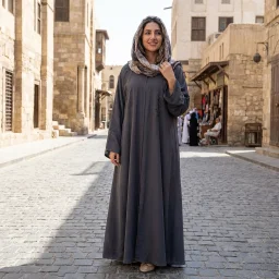 A woman stands on a sunlit stone street wearing a long dark gray abaya and a patterned headscarf. Pale stone buildings, cobblestones, and a few distant people appear in the background.