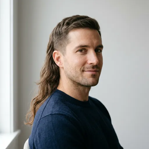 Adult man with a mullet haircut, short on top and sides with long wavy brown hair down the back. He wears a dark navy sweater and faces the camera in soft window light.