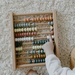 A wooden abacus with colorful beads rests on a cream shag rug while a small child's hand touches one bead near the right side. The scene is photographed from above in soft light.